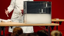 Eine Forscherin steht an einem Tisch. Auf dem Tisch steht ein Experiment mit Kerzen.