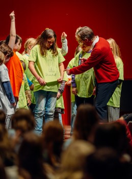 Der Dozent spricht mit einem Kind. Das Kind trägt ein grünes T-Shirt mit der Aufschrift Stickstoff und einer Zeichnung dazu. Im Hintergrund stehen weitere Kinder mit farbigen T-Shirts.