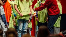 Der Dozent spricht mit einem Kind. Das Kind trägt ein grünes T-Shirt mit der Aufschrift Stickstoff und einer Zeichnung dazu. Im Hintergrund stehen weitere Kinder mit farbigen T-Shirts.