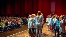 Einige Kinder stehen auf einer Bühne. Im Hintergrund sind viele sitzende Kinder zu sehen.
