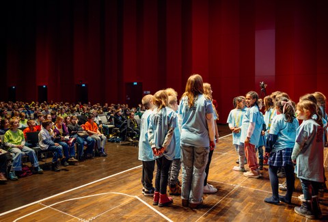 Einige Kinder stehen auf einer Bühne. Im Hintergrund sind viele sitzende Kinder zu sehen.
