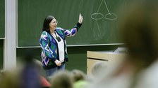 Eine Frau steht vor einer Tafel, auf der eine Kirsche gezeichnet ist. Die Frau erklärt etwas mit den Händen.