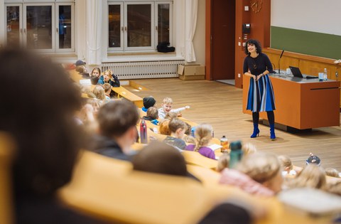 Das Foto zeigt eine Szene in einem Hörsaal. Vor der Tafel steht eine Referentin und spricht zu den Kindern, die in den Reihen sitzen.