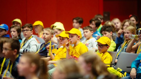 Das Foto zeigt viele Kinder, die im Saal sitzen. Im Zentrum des Fotos sind drei Jungs zu sehen, die das gelbe T-Shirt und Basecap von der Kinder-Uni tragen.  