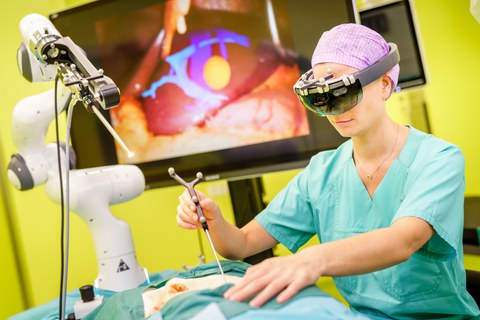 Eine Frau steht an einem OP-Tisch und hat ein OP-Instrument in der Hand. Im Hintergrund ist ein Bildschirm zu sehen. 