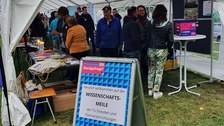 Das Foto zeigt ein großes Festzelt. Im Festzelt sind viele Menschen, die an Tischen stehen oder sich unterhalten. Der Eingang des Festzelts ist offen und davor steht ein Aufsteller mit der Aufschrift Herzlich willkommen auf der Wissenschaftsmeile der TU Dresden und Partnereinrichtungen.