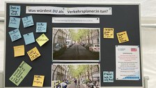 Das Foto zeigt eine Tafel, auf der ganz oben Was würdest DU als Verkehrsplaner:in tun? Darunter steht in kleinerer Schrift Ein Auto benötigt für eine Parkfläche 5 m x 2,50 m. Unter dem Text sind 2 Fotos abgebildet. Das obere Bild zeigte eine Straße mit vielen parkenden Autos. Das untere Bild zeigt die gleiche Straße ohne Autos und mit vielen Menschen. Die Menschen sitzen im Café, gehen spazieren oder beschäftigen sich mit einem Schrank. Neben den beiden Fotos sind Zetteln mit Antworten wie Begrünung, bedachte Straßen, Cafés nur auf einer Seite der Fläche, Rückbau der Versiegelung oder Wasserflächen. Rechts auf der Tafel klebt ein Hinweis auf ein Gewinnspiel.