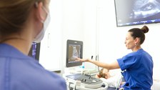 Ärztin führt eine Ultraschalluntersuchung mit dem Sonografiegerät bei einer Person durch, die auf einer Liege liegt; eine weitere Ärztin schaut zu