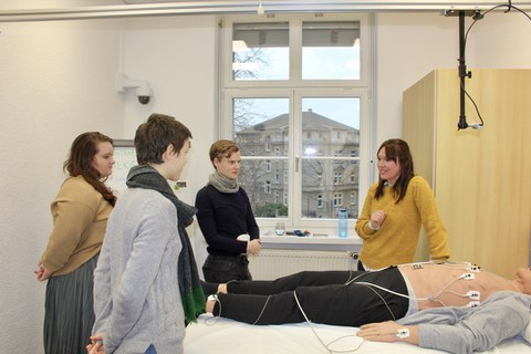 3 Teilnehmende und Dozierende stehen um Dummy mit Elektroden in der Station sensorbasiertes Feedback zur Positionierung von EKG Elektroden