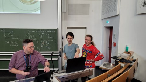 Das Foto zeigt drei Personen in einem Hörsaal bei einer Vorlesung.