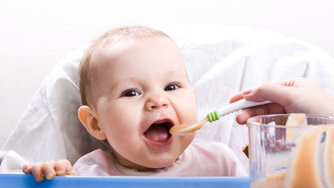 Ein Baby wird mit einem Löffel gefüttert und lacht dabei in die Kamera