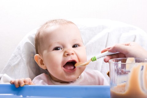 Ein Baby wird mit einem Löffel gefüttert und lacht dabei in die Kamera