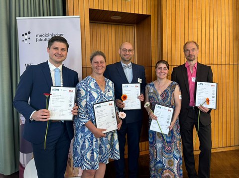 Fünf Personen posieren für das Foto; jede Person hält eine Abschlussurkunde und eine Nelke.