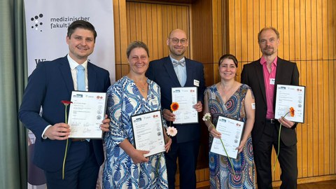Fünf Personen posieren für das Foto; jede Person hält eine Abschlussurkunde und eine Nelke.