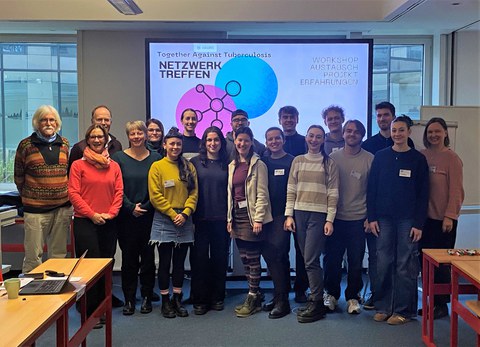 Eine Gruppe Menschen steht in einem Seminarraum vor einem großen Bildschirm mit der Überschrift "Together against Tuberculosis - Netzwerktreffen"