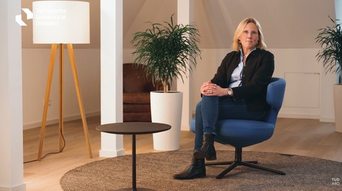 Links stehen Zimmerelemente: Eine Stehlampe, ein Kaffeetisch, ein Pflanzentopf. Rechts: Eine Person sitzt in einem Stuhl 