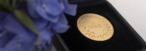 Eine Jung-Medaille in Gold auf schwarzem Hintergrund, im Vordergrund links unfokussiert lila Blumen.