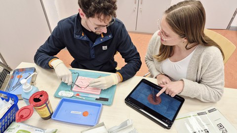 Zwei Menschen sitzen an einem Tisch und üben medizinische Nahttechnik.