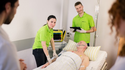 Simulationsperson im Krankenbett und um ihn herum studentische Tutoren und Studierende während einer Lehreinheit