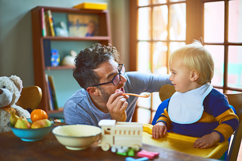 A father feeding his toddler. They are both looking at each other.