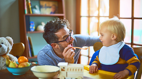 A father feeding his toddler. They are both looking at each other.