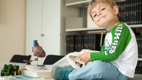 A young boy sits among open medical books, a heart model and a toy tractor on the table in the library, playing with a model of a human skull.