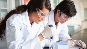 Das Foto zeigt zwei Studierende mit weißen Kitteln und Schutzbrille. Während die Studentin im Vordergrund mit Hilfe einer Pipette Flüssigkeit aus einem Erlenmeyerkolben entnimmt, macht der Student im Hintergrund Notizen auf einem Klemmbrett.