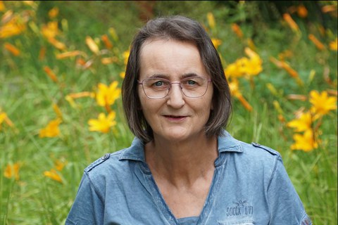 Frau mit Brille und braun-grauen Haaren in blauem Oberteil vor einem Blumen-Hintergrund