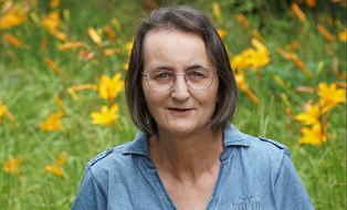 Frau mit Brille und braun-grauen Haaren in blauem Oberteil vor einem Blumen-Hintergrund