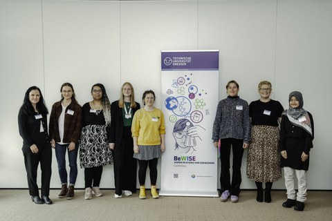 Sieben Frauen stehen neben einem Poster auf dem eine Frau mit Erlenmeyerkolben abgebildet ist, sowie das TU Dresden logo und der Schriftzug BeWise