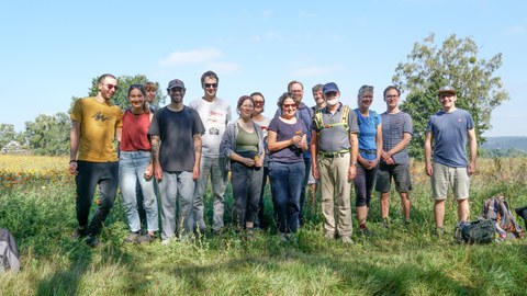 Sechs Frauen und acht Männer stehen an einem sonnigen Tag nebeneinander auf einem Feld.