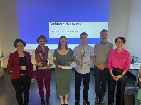 Zwei Frauen und ein Mann stehen neben zweiFrauen und einem Mann, die jeweils eine gelbe Rose und ein Papier in der Hand halten. Im Hintergrunf ist eine Präsentation zu sehen auf welcher Fachbereich Chemie steht.