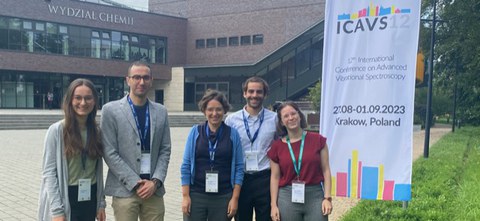 Drei Frauen und ein Mann stehen vor einem Gebäude und einem banner auf dem ICAVS12 steht. Die Menschen tragen Ausweise um den Hals.