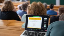 Im Hörsaal sitzen viele Menschen. An einem Platz steht ein aufgeklappter Laptop und zeigt den Titel des Vortrags.