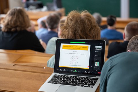 Im Hörsaal sitzen viele Menschen. An einem Platz steht ein aufgeklappter Laptop und zeigt den Titel des Vortrags.