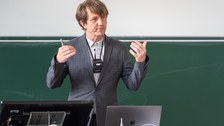 Ein Mann steht in einem Hörsaal mit dem Rücken zur Tafel.