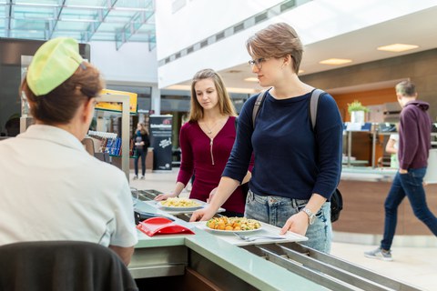 Das Foto zeigt zwei Studierende in der Mensa. Sie stehen gerade an der Kasse und bezahlen ihr Essen.