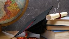 Das Foto zeigt einen verschiedene Gegenstände, die nebeneinander auf einem Tisch liegen. Unter anderem erkennt man einen Globus, einen Doktorhut, ein Diplom und mehrere Bücher.