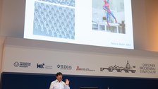 Andre Geim hebt die Hand. Im Hintergrund sieht man eine Darstellug von Spiderman, der die gleiche Geste macht.
