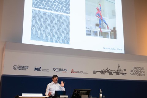 Andre Geim hebt die Hand. Im Hintergrund sieht man eine Darstellug von Spiderman, der die gleiche Geste macht.