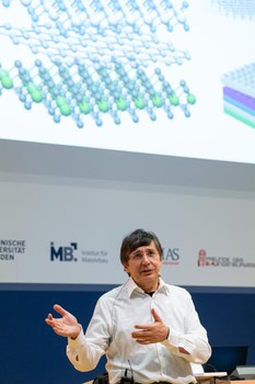 Andre Geim hebt erklärend die Hände. Im HIntergrund die Power Point Präsentation.