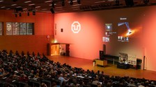 Foto von hinten über die Köpfe der Zuschauenden hinweg. Vorne sieht man die Powerpoint von Reinhard Genzel auf der er und eine Darstellung eines schwarzen Lochs ist
