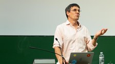 Physik Nobelpreisträger Didier Queloz am Pult vom Audimax der TU Dresden. Unter den vier Stickern auf seinem MacBook ist ein blauer mit einem Fisch. Um diesen steht: "JA zur Biodiversität" 