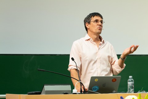 Physik Nobelpreisträger Didier Queloz am Pult vom Audimax der TU Dresden. Unter den vier Stickern auf seinem MacBook ist ein blauer mit einem Fisch. Um diesen steht: "JA zur Biodiversität" 