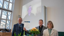 Drei Personen stehen im Vordergrund mit einer Urkunde und Blumen. Im Hintergrund ist eine Person an der Leinwand per Video zugeschalten.