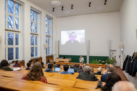 Hörsaalansicht von hinten nach vorn. Auf der Leinwand ist ein Mann zu sehen.
