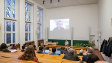 Hörsaalansicht von hinten nach vorn. Auf der Leinwand ist ein Mann zu sehen.