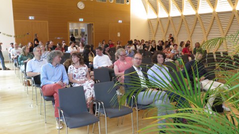 In einem großen Saal sitzen viele Menschen. in der vordersten Reihe sind 2 Plätze frei