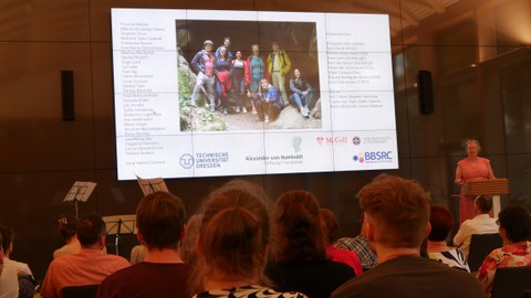 Auf dem Bildschirm im großen Bild sieht man ein Gruppenbild einer Forschergruppe 