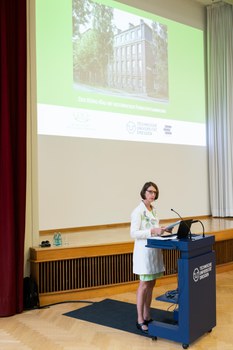 Frau in gelben Blazer und grünem Kleid steht am Pult. Hinter ihr wird eine Powerpoint mit einem Bild vom König Bau